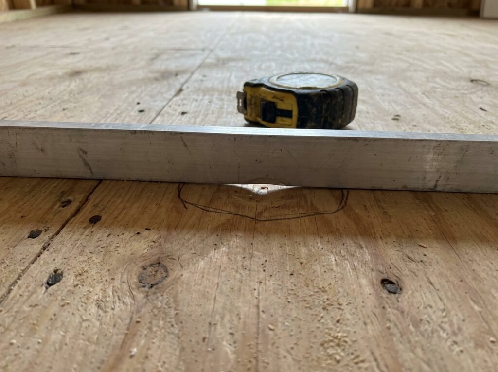 Straightedge on a plywood subfloor revealing a low spot outlined in pencil