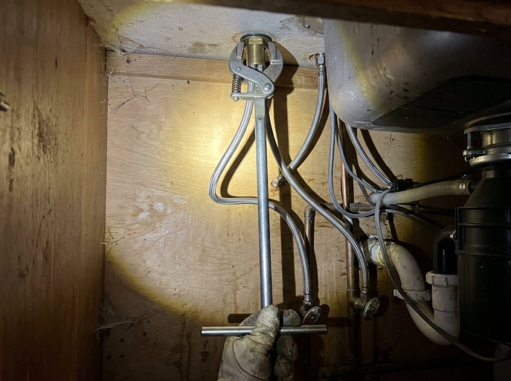 Basin wrench loosening a faucet mounting nut under a kitchen sink with braided supply lines visible