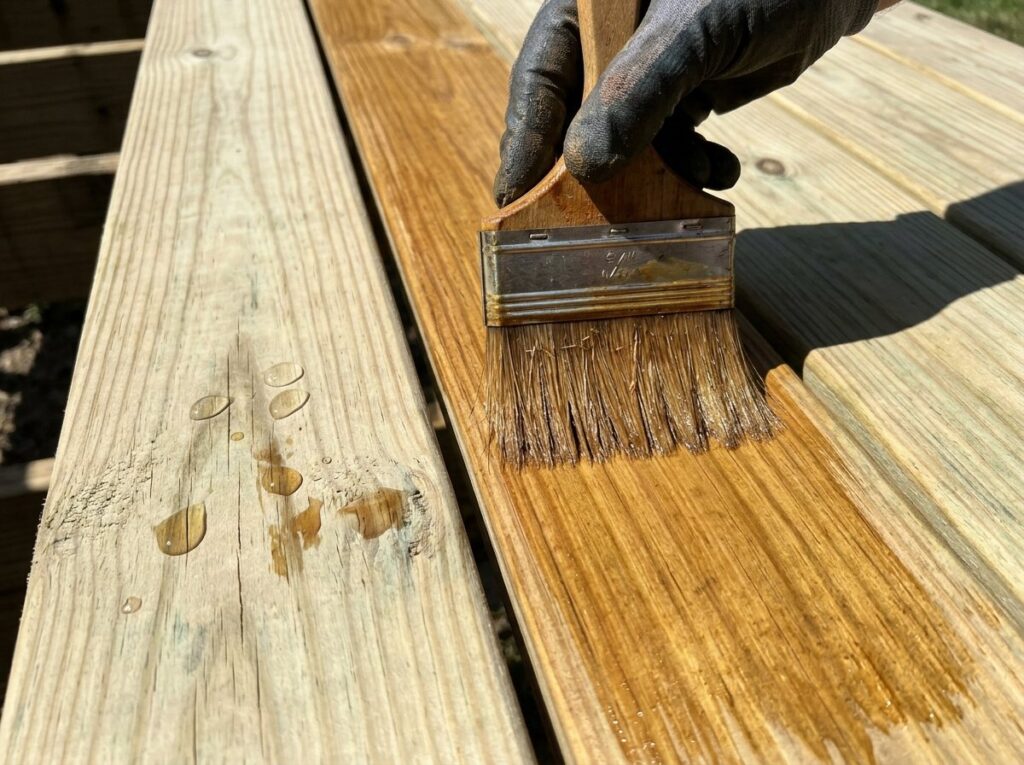Gloved hand back-brushing semi-transparent stain into clean deck boards along the grain